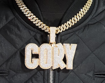 May include: A gold-toned cuban link chain necklace featuring a large, block-letter pendant encrusted with clear stones that spells out the name CORY. The pendant is attached to the chain by a rectangular, stone-covered bail.