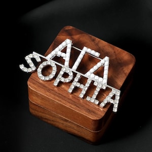 Pode incluir: Broche prateado com o nome "SOPHIA" e as letras gregas "ΑΓΑ", incrustado com brilhantes pedras de strass. O broche é exibido em uma pequena caixa de madeira quadrada, sobre um fundo escuro.