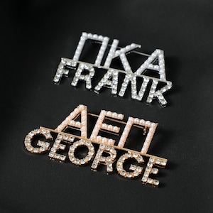 Op de afbeelding: Twee decoratieve broches met tekst. De bovenste broche is zilverkleurig met de tekst "PIKA FRANK" uitgespeld in witte imitatieparels. De onderste broche is goudkleurig met de tekst "AEII GEORGE" uitgespeld in heldere kristallen en witte imitatieparels.
