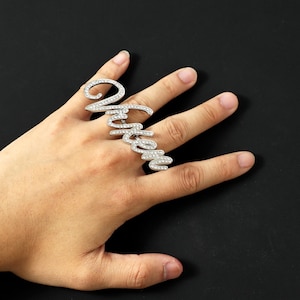 Peut inclure: Bague argentée avec l'inscription "Queen" en écriture cursive, incrustée de petits cristaux transparents. La bague est présentée sur une main, sur fond noir.