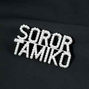 Può includere: Spilla argentata con le parole "SOROR TAMIKO" scritte in maiuscolo. Le lettere sono delineate da piccole perle bianche rotonde. La spilla è posta su uno sfondo nero.