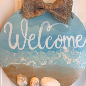 May include: Circular wooden welcome sign with a painted beach scene. The sign has the word "Welcome" in white script, a light blue sky, and a brown sand area with three seashells. A burlap bow is attached at the top.