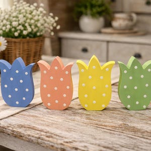 May include: Four wooden tulip decorations in blue, peach, yellow, and green, each with white polka dots. They are arranged on a wooden table with a striped runner, with baskets of flowers in the background.