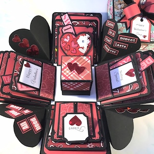 May include: A black and red explosion box with multiple layers. Each layer has a different design with hearts, plaid, and lace. The box is decorated with the words "Love is a moment that lasts forever", "Happy Valentine's", and "Nothing Special".