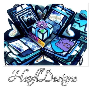 May include: A blue and black explosion box with a small gift box on top. The gift box is decorated with a blue and white checkered pattern and a purple flower. The explosion box has various compartments with different designs and text, including "Mazel Tov" and "Bat Mitzvah".