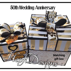 May include: Two matching gift boxes for a 50th wedding anniversary. The boxes are white with gold stripes and black trim. Each box has a large bow with gold and black accents. One box has a heart and the words "Best Wishes".