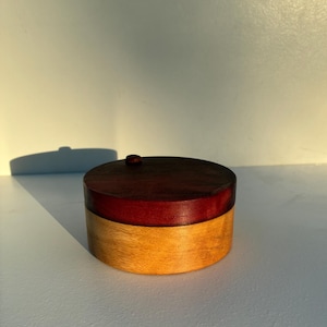 Könnte beinhalten: Eine zylindrische Holzschatulle mit zweifarbigem Design, bestehend aus einem dunkel rotbraunen Deckel und einem helleren goldbraunen Unterteil. Der Deckel verfügt über einen kleinen, runden Holzknopf auf der Oberseite und ist kreisförmig.