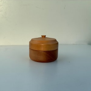 Puede incluir: Una pequeña caja redonda de madera con tapa. La caja es de dos tonos de madera, con un color más claro para la tapa y un color más oscuro para la base. La tapa tiene un pequeño pomo.