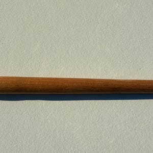 Può includere: Un utensile in legno liscio color marrone chiaro con un corpo cilindrico che si assottiglia verso un'estremità. L'estremità opposta presenta un piccolo pomello arrotondato seguito da un dettaglio svasato a forma di campana. Venature visibili.