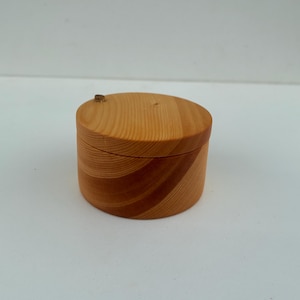 May include: A small, cylindrical wooden box with a lid, featuring a natural light brown wood grain pattern. The container has a smooth, polished finish and a circular shape, showcasing the distinct rings and textures of the wood grain throughout.