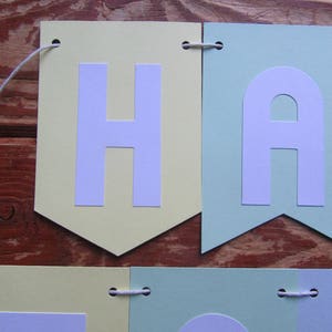 Pode incluir: Uma faixa de cores pastel com a palavra "Happy" escrita em letras brancas. Cada letra est&aacute; em um peda&ccedil;o de papel&atilde;o separado, com cores alternadas de amarelo claro e verde claro. A faixa &eacute; amarrada com um barbante branco.