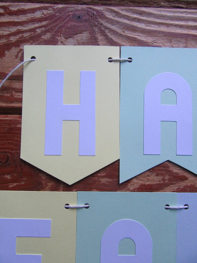 Pode incluir: Uma faixa de cores pastel com a palavra "Happy" escrita em letras brancas. Cada letra est&aacute; em um peda&ccedil;o de papel&atilde;o separado, com cores alternadas de amarelo claro e verde claro. A faixa &eacute; amarrada com um barbante branco.