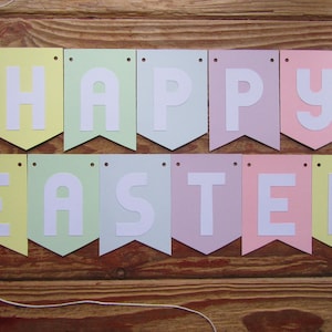 Pode incluir: Uma faixa de cores pastel com as palavras "Happy Easter" em letras brancas. A faixa &eacute; feita de papel e tem um cord&atilde;o amarrado.