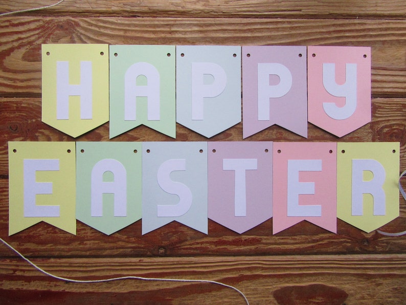 Pode incluir: Uma faixa de cores pastel com as palavras "Happy Easter" em letras brancas. A faixa &eacute; feita de papel e tem um cord&atilde;o amarrado.