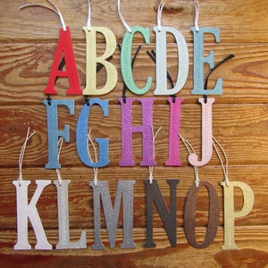 May include: A set of 16 colourful glitter letters, A through P, on a wooden background. Each letter is attached to a string.