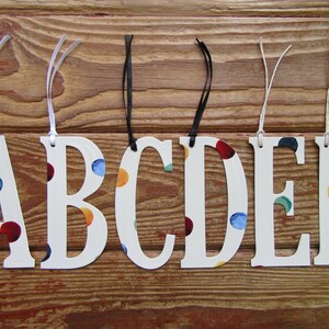 Pode incluir: Seis letras de madeira brancas, A, B, C, D, E e F, com um padrão de bolinhas coloridas. Cada letra tem um cordão para pendurar.