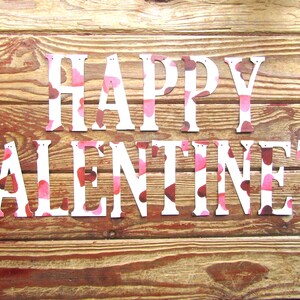 Pode incluir: Letras brancas que soletram "Happy Valentines" em um fundo de madeira. As letras são decoradas com um padrão rosa e marrom.
