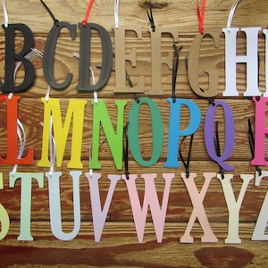 Pode incluir: Um conjunto de 26 letras de madeira, cada uma pintada de uma cor diferente, penduradas em um cordão. As letras estão em ordem alfabética, começando com A e terminando com Z. As letras são todas maiúsculas e têm aproximadamente 5 cm de altura.