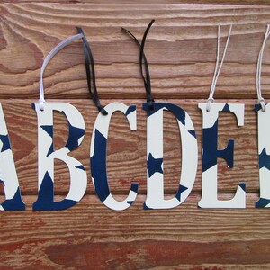 Pode incluir: Seis letras de madeira brancas, de A a F, com um padrão de estrelas azul e branco. Cada letra tem um cordão para pendurar.