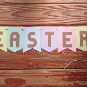 Pode incluir: Uma faixa de cores pastel com a palavra "EASTER" escrita em letras marrons. A faixa est&aacute; amarrada com um barbante branco e est&aacute; pendurada em uma superf&iacute;cie de madeira.