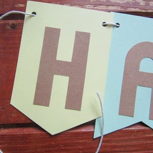 Pode incluir: Uma faixa de papel verde claro e azul claro com a palavra "Happy" em letras marrons. A faixa &eacute; amarrada com um barbante branco.