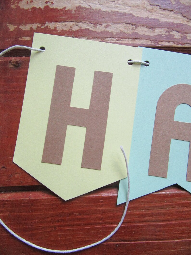 Pode incluir: Uma faixa de papel verde claro e azul claro com a palavra "Happy" em letras marrons. A faixa &eacute; amarrada com um barbante branco.
