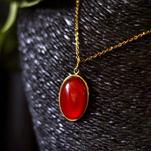 Può includere: Una collana dorata con un pendente a forma di pietra rossa ovale. Il pendente è incastonato in una cornice dorata e appeso a una sottile catena d'oro. Lo sfondo è una superficie scura e strutturata.