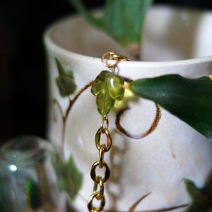 May include: A gold chain with a leaf-shaped charm hangs near a white ceramic pot with green leaf designs. The chain is made of small, linked circles. A green plant leaf is visible.
