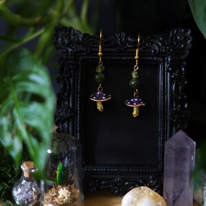 Peut inclure: Boucles d'oreilles en forme de champignon avec des chapeaux violets à pois blancs, des tiges dorées et des perles vertes. Les boucles d'oreilles sont présentées devant un cadre noir, avec un petit bocal en verre et des cristaux en arrière-plan.