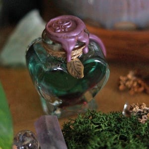 Peut inclure: Une bouteille en verre en forme de cœur remplie d'un liquide vert translucide. La bouteille est scellée avec un sceau en cire violette et attachée avec de la ficelle ornée d'une petite breloque en métal en forme de feuille.