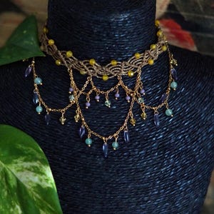 Peut inclure: Un collier ras-du-cou en macramé composé d'une base en cordon marron tressé avec des perles jaunes incrustées. Il comporte plusieurs chaînes dorées superposées, ornées de petites perles pendantes violettes, bleu clair et vert pâle.