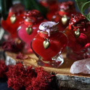 Op de afbeelding: Verschillende hartvormige glazen flessen gevuld met een helderrode vloeistof. Elke fles heeft een gouden hartvormige bedel en een decoratieve dop. De flessen staan op een houten oppervlak met rood mos en een rozenkwartskristal.