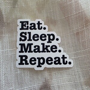 Könnte beinhalten: Weißer Aufkleber mit schwarzem Text: "Eat. Sleep. Make. Repeat." Der Aufkleber hat eine rechteckige Form mit abgerundeten Ecken. Der Hintergrund ist ein strukturiertes, hellbeige farbenes Gewebe.