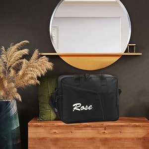 Könnte beinhalten: Schwarze Reisetasche mit einem weißen gestickten Namen "Rose" auf der Vorderseite. Die Tasche liegt auf einem Holzregal mit einem runden Spiegel darüber.