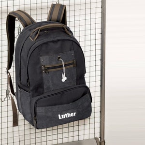 以下が含まれることがあります： 黒色のバックパック。茶色のアクセントがあり、金属製の格子に掛けられています。「Luther」という名前が白でプリントされたフロントポケットがあります。上部のハンドル、調節可能なストラップ、ジッパー付きのフロントポケットが付いています。白いイヤホンが見えます。