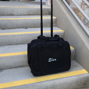 Könnte beinhalten: Schwarze Rollreisetasche mit Griff und Rollen. Die Tasche hat den Namen "Elsa" auf der Vorderseite gestickt.
