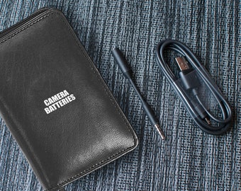 May include: A black leather case with the text "CAMERA BATTERIES" printed on it, a black USB cable, and a black audio cable.
