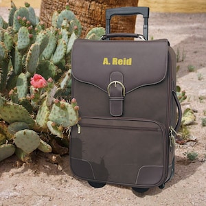 May include: A brown rolling suitcase with a leather-like finish and a gold buckle. The suitcase has a name tag that reads "A. Reid".