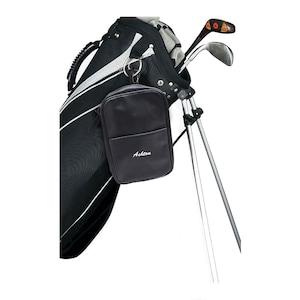 May include: A black golf bag with a silver zipper and a black leather pouch with the word "Ashton" embroidered on it.