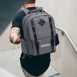 May include: Gray backpack with black leather accents, buckles, and straps. Features a top handle, side pocket, and the name "SAMMY" printed on the front. The backpack is being carried up a set of stairs.