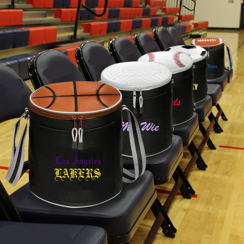 Puede incluir: Una fila de bolsas aislantes negras y marrones con diferentes logotipos de equipos deportivos. Las bolsas est&aacute;n alineadas en sillas negras en un gimnasio. Los logotipos incluyen los Los Angeles Lakers, una pelota de b&eacute;isbol, una pelota de golf y una pelota de f&uacute;tbol.