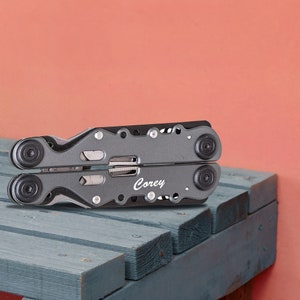 May include: A gray multi-tool with a black rubber handle and the name "Corey" engraved on the side. The multi-tool is sitting on a gray wooden surface.