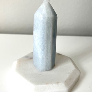 Può includere: Una punta di cristallo blu chiaro con una cima arrotondata, appoggiata su una base di marmo bianco. La punta di cristallo ha una forma esagonale e una superficie liscia e lucida.