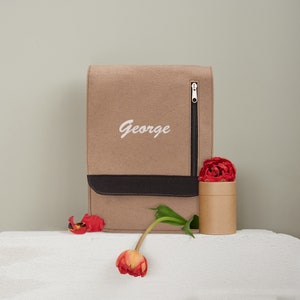 May include: A brown felt laptop bag with a black leather trim and a zipper. The bag has the name "George" embroidered on the front in white. A red tulip is next to the bag.