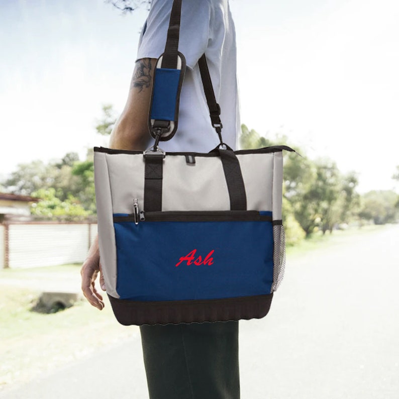 Może przedstawiać: Szara i niebieska torba typu tote z czarnym paskiem na ramię i czarnym uchwytem. Torba ma zamek błyskawiczny i siatkową kieszeń z boku. Torba jest haftowana imieniem "Ash" na czerwono.