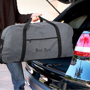 May include: A gray duffel bag with black straps and a zippered pocket. The bag has the words "Best Dad" embroidered on the front in black.