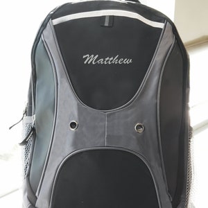 May include: Black and gray backpack with a white stripe and the name "Matthew" embroidered on the front.