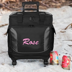 May include: Black rolling cooler bag with a pink "Rose" embroidered on the front. The cooler bag has two wheels and a handle. Two cans of soda are in the foreground.