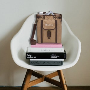 Könnte beinhalten: Eine braune Canvas-Tragetasche mit einem braunen Lederriemen und einer braunen Lederklappe mit einer Schnalle. Die Tasche liegt auf einem Stapel Bücher. Die Tasche hat das Wort "Robinsons" auf der Vorderseite gestickt.