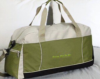 May include: A green and beige travel duffel bag with a black zipper and a black bottom. The bag has a shoulder strap and two top handles. The bag has the text "Blessings Wait For You" embroidered on the front.